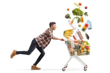Alışveriş arabasıyla koşan mutlu genç bir adam ve beyaz arka planda izole edilmiş düşen yiyecekler.