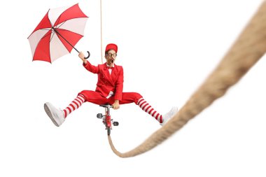Kırmızı takım elbiseli bir şovmen ip üzerinde küçük bir bisiklet sürüyor ve elinde beyaz arka planda izole edilmiş bir şemsiye tutuyor.