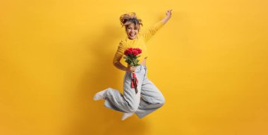 Mutlu Asyalı kadın elinde bir demet kırmızı gülle sarı arka plana atlıyor. 