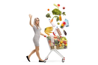 Düşen gıda ürünleriyle alışveriş arabasını iten ve beyaz arka planda izole edilmiş kameraya gülümseyen bir kadın.