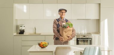 Mutfakta duran ve elinde poşet dolusu yiyecek tutan yaşlı bir adam.