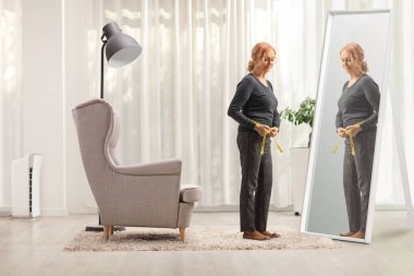 Düşünceli, olgun bir kadının evde aynanın önünde belini ölçerken tam boy fotoğrafı.