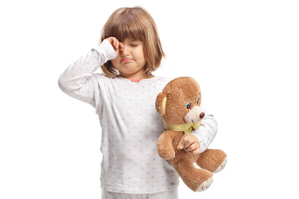 Girl in pajamas holding a teddy bear and rubbing eyes isolated on white background