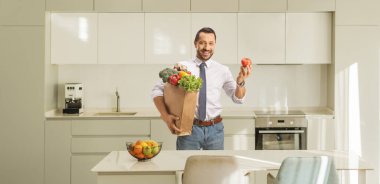 Mutfakta bakkal poşeti ve domates tutan neşeli genç bir adam.