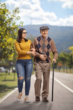 Genç bir kadın parkta bastonla yürüyen yaşlı bir adama yardım ediyor.