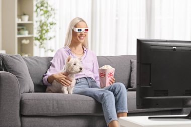 3D gözlüklü bir kadın bir kanepede oturuyor ve bir köpekle televizyon izliyor. 