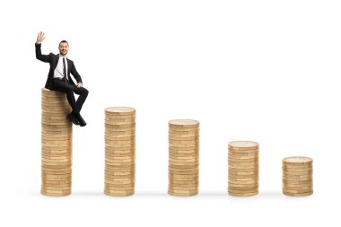 İşadamı en yüksek madeni para yığınında oturuyor ve beyaz arka planda izole bir şekilde el sallıyor.