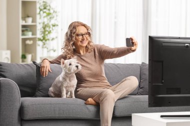 Kanepede oturan bir kadın, elinde bir teriyer köpeğiyle akıllı telefon kamerasıyla selfie çekiyor.