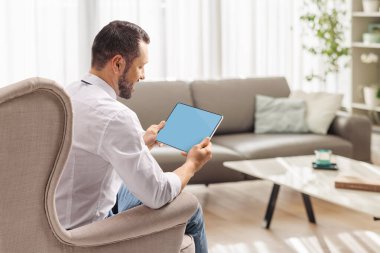 Evdeki koltukta oturan ve oturma odasında dijital tablet kullanan bir adam.