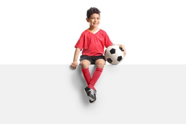 Futbol formalı Asyalı bir çocuk beyaz arka planda boş bir panelde oturuyor.