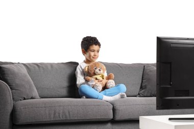 Kanepede oturan bir çocuk oyuncak ayıyı tutuyor ve beyaz arka planda izole bir şekilde televizyon izliyor.