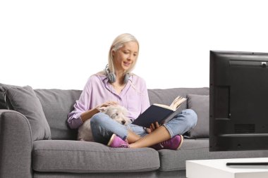 Kanepede bir köpekle oturan ve beyaz arka planda izole bir şekilde televizyon karşısında kitap okuyan bir kadın.
