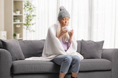 Genç bir kadın battaniyeye sarılmış kanepede oturuyor ve evde sıcak çay içiyor.