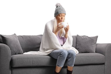 Battaniyeye sarılı genç bir kadın kanepeye oturup beyaz arka planda izole edilmiş çayı içiyor.