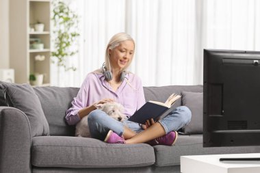 Kanepede oturan bir kadın batılı bir teriyeri köpeğini okşuyor ve kitap okuyor.