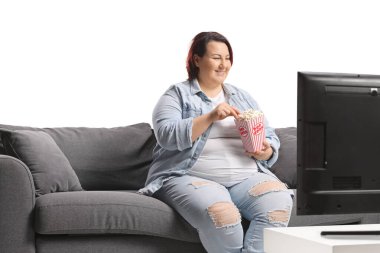 Kanepede oturan, televizyon izleyen ve beyaz arka planda izole edilmiş patlamış mısır yiyen bir kadın.