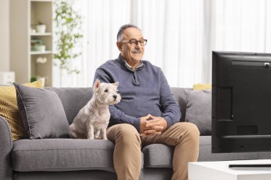 Kanepede oturan ve köpeğiyle televizyon izleyen gülümseyen olgun bir adam.  
