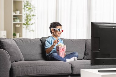 Kanepede oturan bir çocuk 3 boyutlu gözlüklü televizyon izliyor ve patlamış mısır yiyor. 