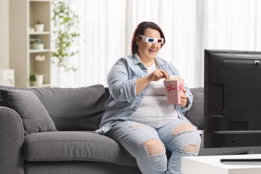 Kanepede oturan bir kadın 3 boyutlu gözlüklü televizyon izliyor ve patlamış mısır yiyor. 