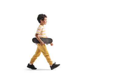 Beyaz arka planda yürüyen ve kaykay taşıyan bir çocuğun tam boy profil çekimi.