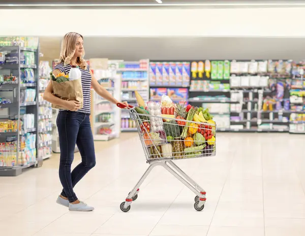 Tam boy profil, süpermarkette elinde alışveriş arabası ve market çantasıyla duran genç bir kadını vurdu.