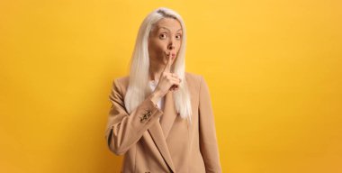 Sarışın bir kadın sarı bir arka planda sessizce el sallıyor.