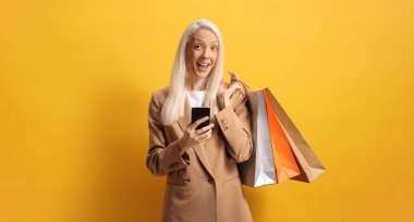 Sarı arka planda akıllı telefon ve alışveriş torbaları tutan mutlu kadın.