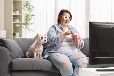 Bir kutu patlamış mısır ve 3D gözlüklü şişman bir kadın bir kanepede oturmuş televizyon seyrediyor.