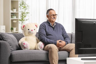 Elinde oyuncak ayıyla kanepede oturan ve evde televizyon izleyen olgun bir adam.