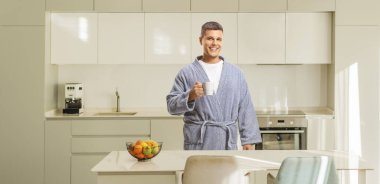 Bornozlu genç adam mutfakta bardak tutuyor.