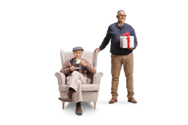 Mature man holding a present and standing to a senior seated in an armchair isolated on white background