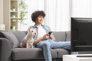 Köpekli genç adam televizyon izliyor ve elinde cep telefonu var.