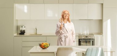 Mutfakta elinde bardak tutan bornozlu olgun bir kadın.