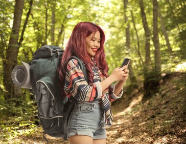 Ormanda duran sırt çantalı ve akıllı telefon kullanan genç bir bayan yürüyüşçü.