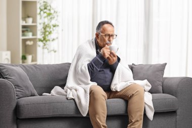 Battaniyeye sarılmış, kanepeye oturmuş çay içen olgun bir adam. 