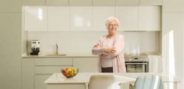 Mutfakta gülümseyen ve kollarını kavuşturarak poz veren yaşlı bir kadın. 