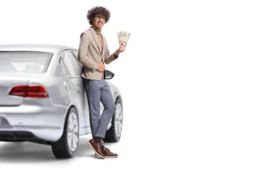 Genç adam arabaya yaslanıyor ve beyaz arka planda izole edilmiş yığınla para tutuyor.