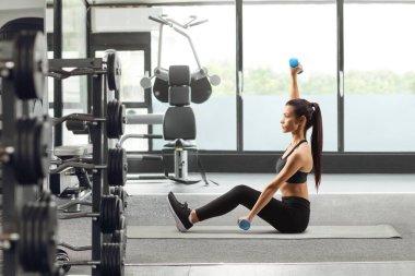 Spor salonundaki paspasın üzerinde dambıllarla egzersiz yapan genç bir kadın.