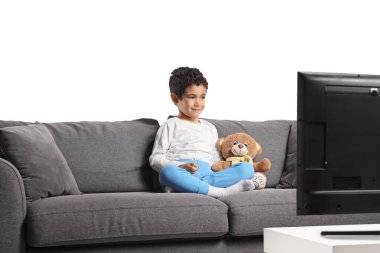 Pijamalı küçük çocuk bir kanepede oyuncak ayıyla oturuyor ve beyaz arka planda izole bir şekilde televizyon izliyor.