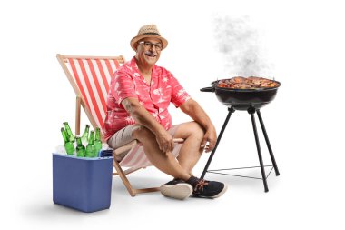 Güverte sandalyesinde oturan bir adam, buzdolabında biralar ve beyaz arka planda ızgara et yiyen bir adam.