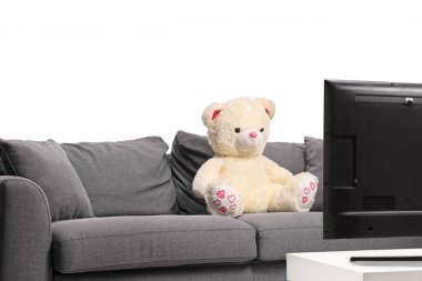Beyaz arka planda izole edilmiş bir televizyon ekranının önündeki kanepede oturan oyuncak ayı.