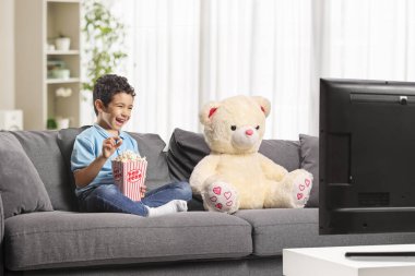 Çocuk patlamış mısır yiyor ve oyuncak ayısıyla televizyon seyrediyor.