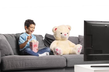 Mutlu çocuk patlamış mısır yiyor ve beyaz arka planda izole edilmiş oyuncak ayısıyla televizyon izliyor.