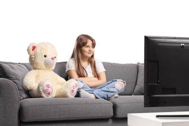 Kanepede oturan ve beyaz arka planda izole televizyon izleyen oyuncak ayılı bir kız.