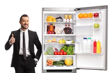 Takım elbiseli genç adam açık bir buzdolabına yaslanıyor ve parmakları beyaz arka planda izole edilmiş.