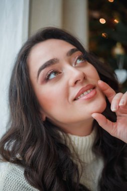 Açık renk örgü kazaklı mutlu, siyah saçlı kıvırcık kadın Noel ağacının yanında hediyeler hazırlıyor. Şık bir model kış tatilinde online alışveriş yapıyor. Yeni yıl. Konforlu ev.
