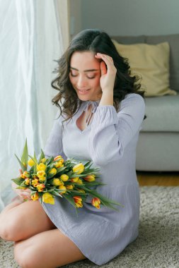 Spring portrait of beautiful dark haired curly woman in lilac dress with large color bouquet of tulips at home. Emotional model holds flowers in her hands. Womens Day. Present. Mothers Day.
