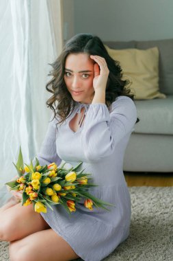 Spring portrait of beautiful dark haired curly woman in lilac dress with large color bouquet of tulips at home. Emotional model holds flowers in her hands. Womens Day. Present. Mothers Day.