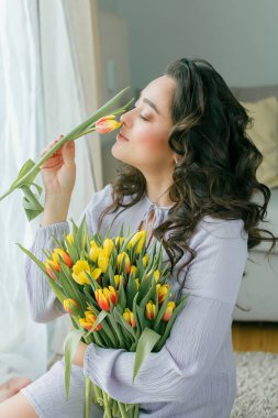 Spring portrait of beautiful dark haired curly woman in lilac dress with large color bouquet of tulips at home. Emotional model holds flowers in her hands. Womens Day. Present. Mothers Day.