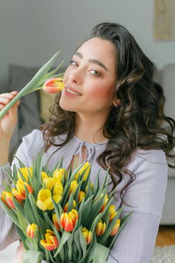 Spring portrait of beautiful dark haired curly woman in lilac dress with large color bouquet of tulips at home. Emotional model holds flowers in her hands. Womens Day. Present. Mothers Day.
